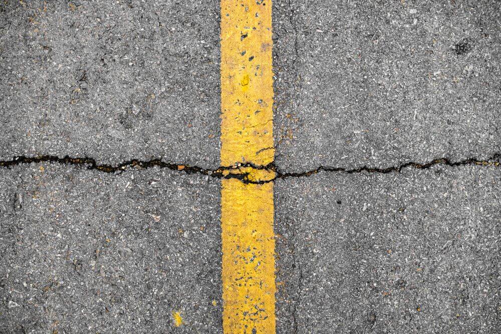 Closeup photo of an asphalt road with a yellow line and a big crack going across it.