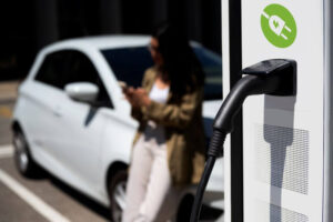 electric car charging station with a blurred out woman leaning on her car in the background waiting for it to charge